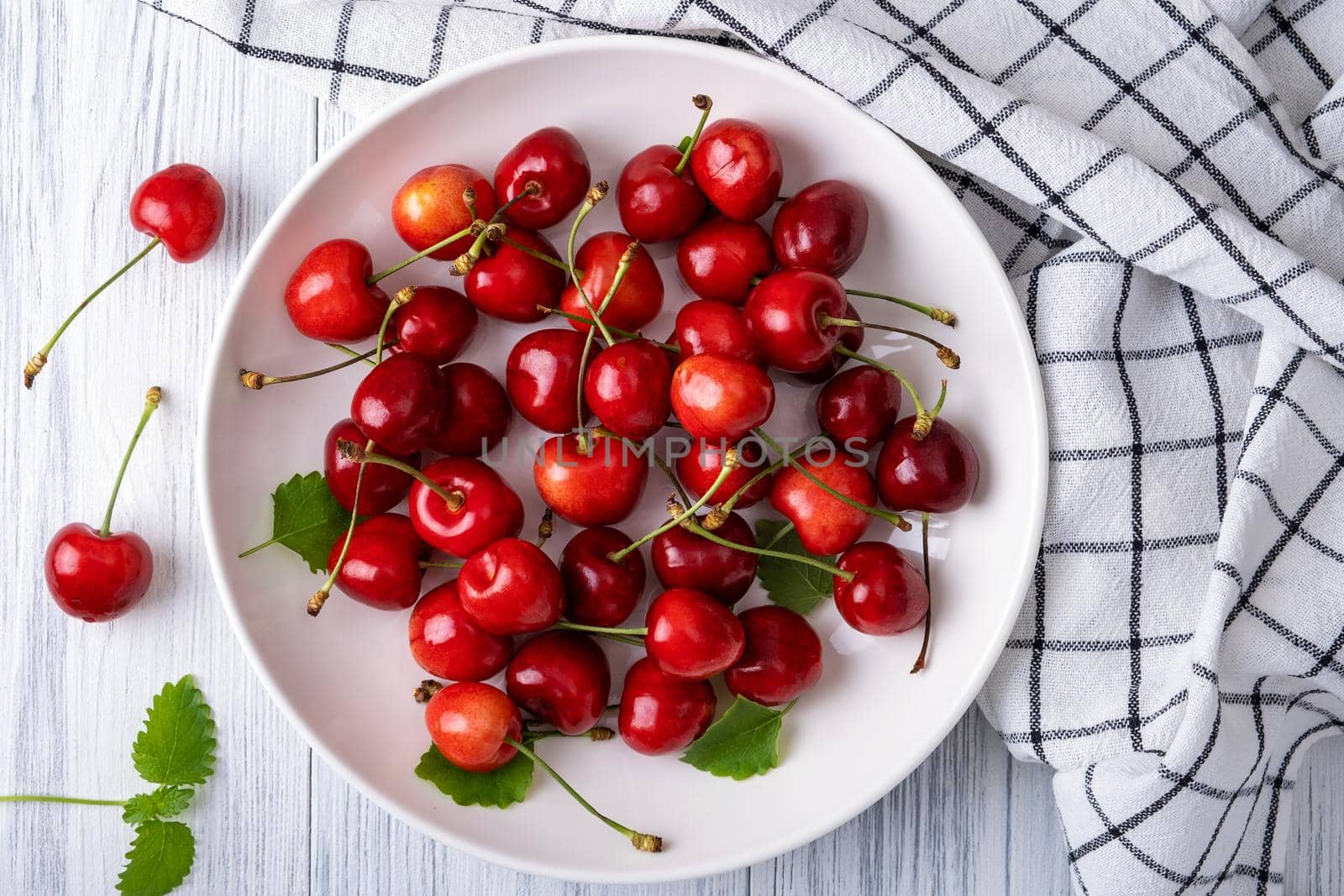 Ripe juicy sweet cherry lies on a white porcelain plate on a light wooden surface. Summer healthy food. Selective focus. View from above.