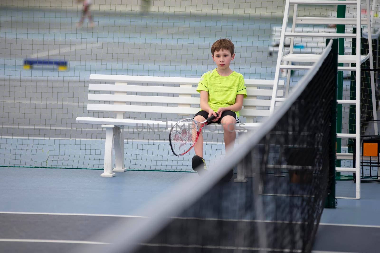 Sad boy sits on a bench at a tennis competition. The child plays tennis.