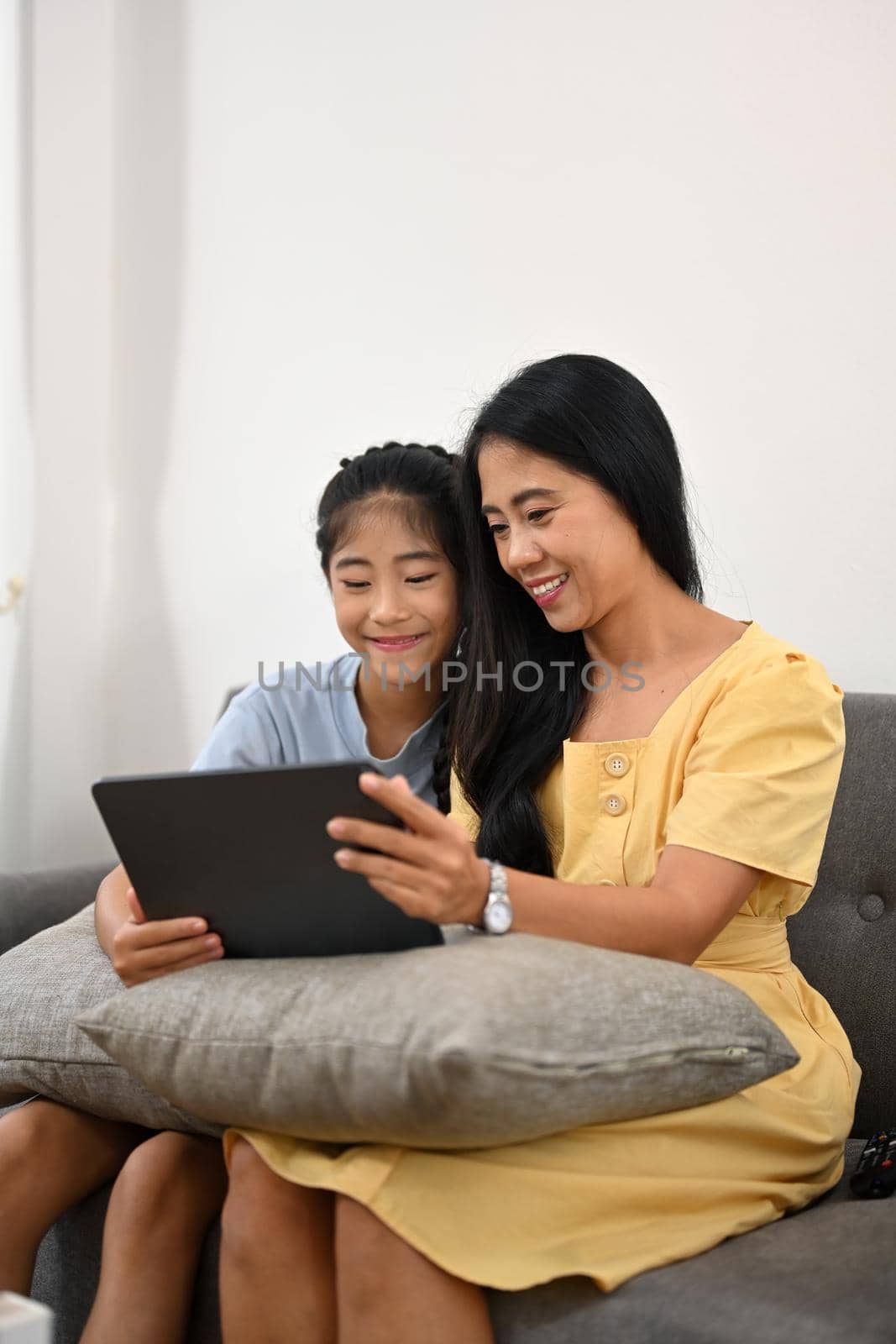 Asian mom and daughter having fun browsing internet on digital tablet together on sofa.