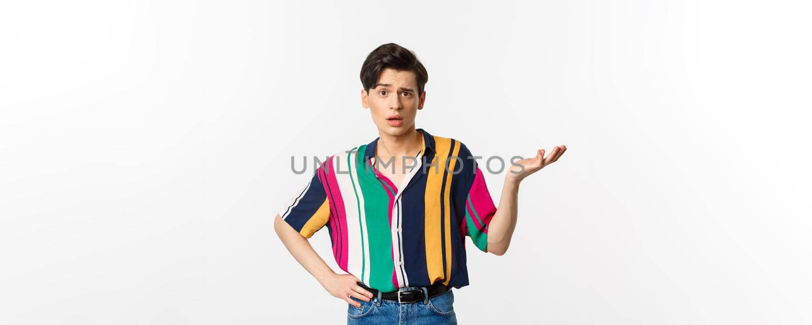 Confused gay man asking what happened, raising hand aside and looking puzzled at camera, standing over white background.