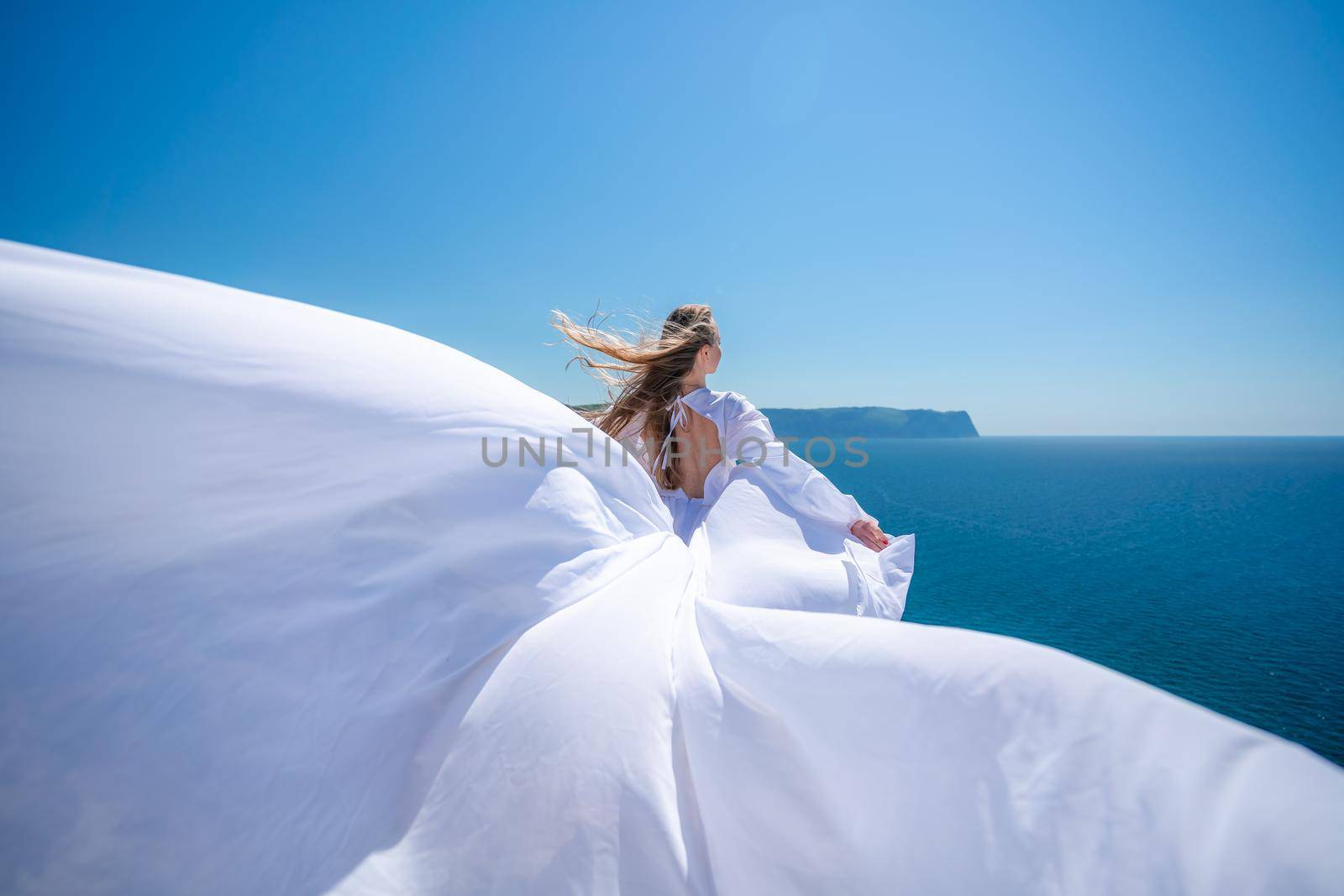 Blonde with long hair on a sunny seashore in a white flowing dress, rear view, silk fabric waving in the wind. Against the backdrop of the blue sky and mountains on the seashore