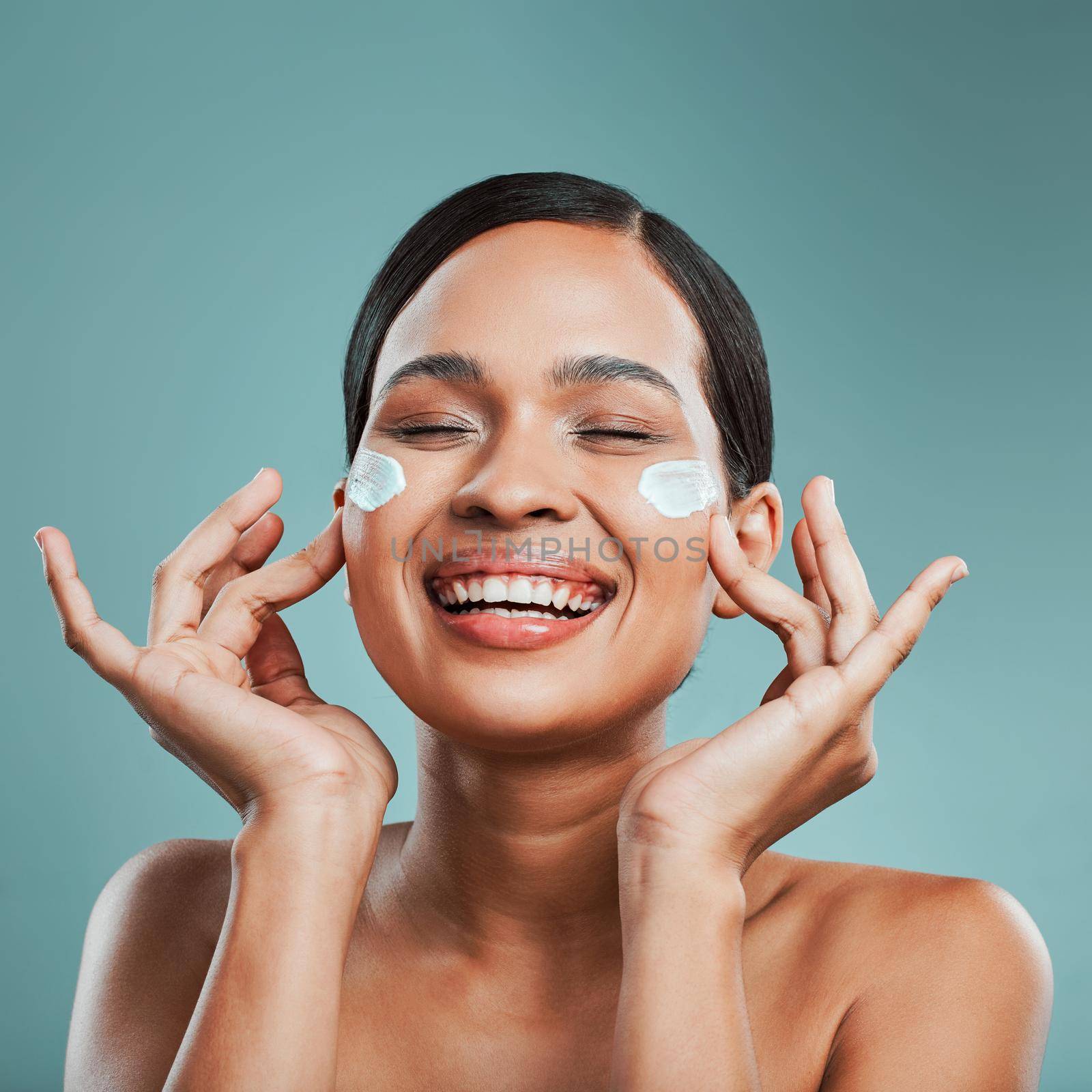 An attractive young mixed race woman posing with cream moisturiser on her face against a green studio background. Beautiful hispanic using a skin products to keep her face clean and healthy by YuriArcurs