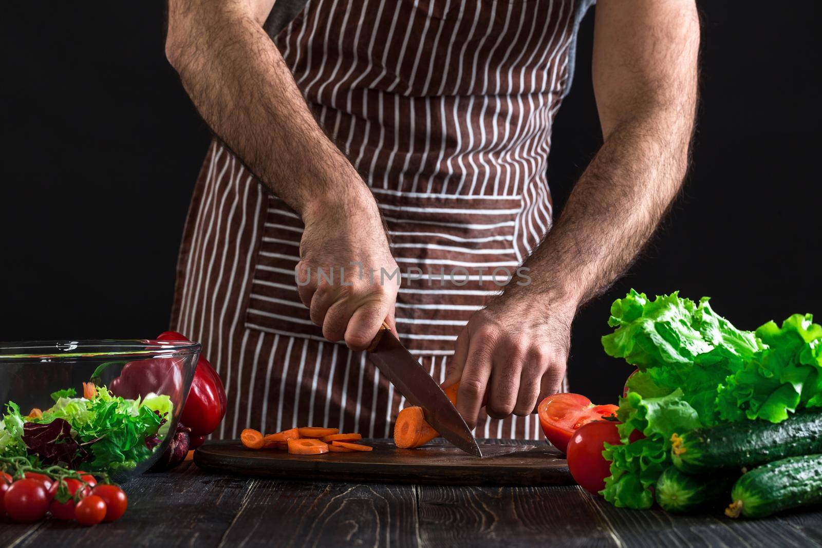 Young home cook man in apron slicing carrot with kitchen knife. Men's hands cut the carrot to make a salad on black background. Healthy food concept