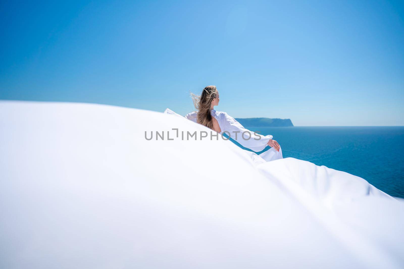 Blonde with long hair on a sunny seashore in a white flowing dress, rear view, silk fabric waving in the wind. Against the backdrop of the blue sky and mountains on the seashore