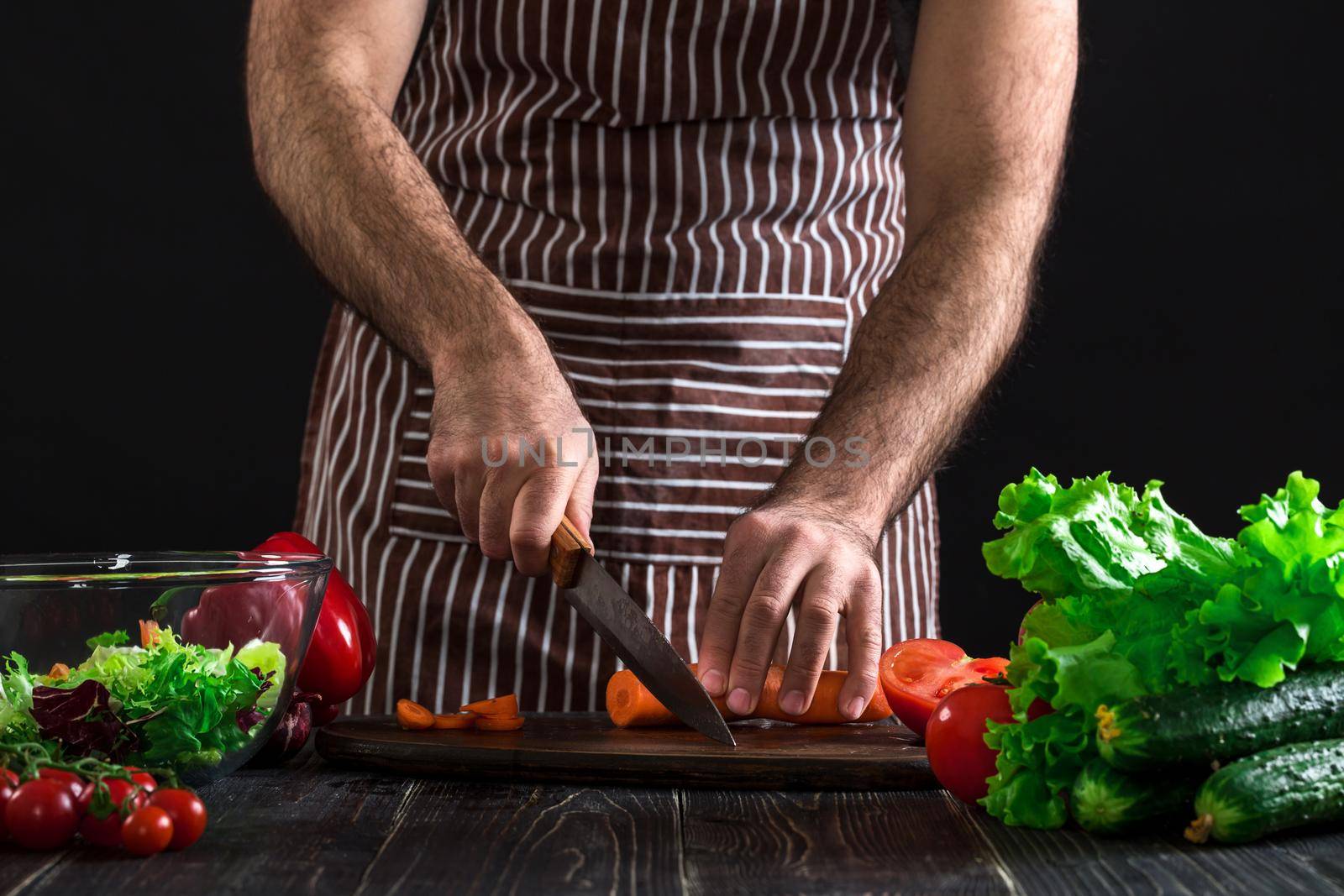 Young home cook man in apron slicing carrot with kitchen knife. Men's hands cut the carrot to make a salad on black background. Healthy food concept