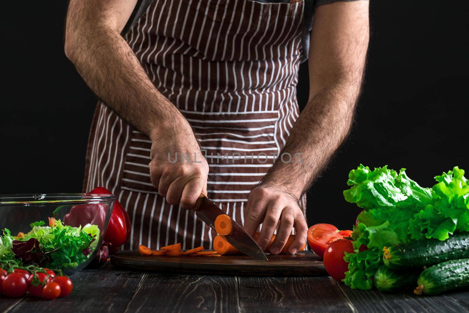 Young home cook man in apron slicing carrot with kitchen knife. Men's hands cut the carrot to make a salad on black background. Healthy food concept