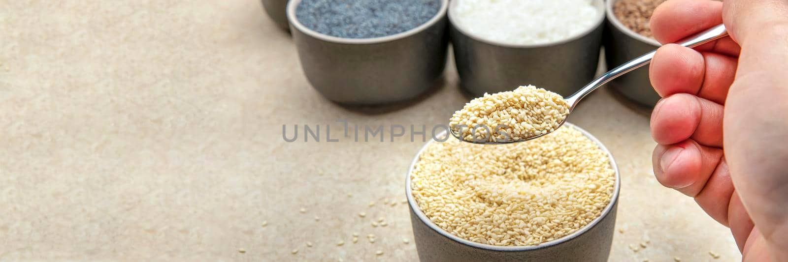 White sesame seeds, sesame seeds in a black saucer on a background of various seeds. Male hand sprinkles sesame seeds with a spoon.