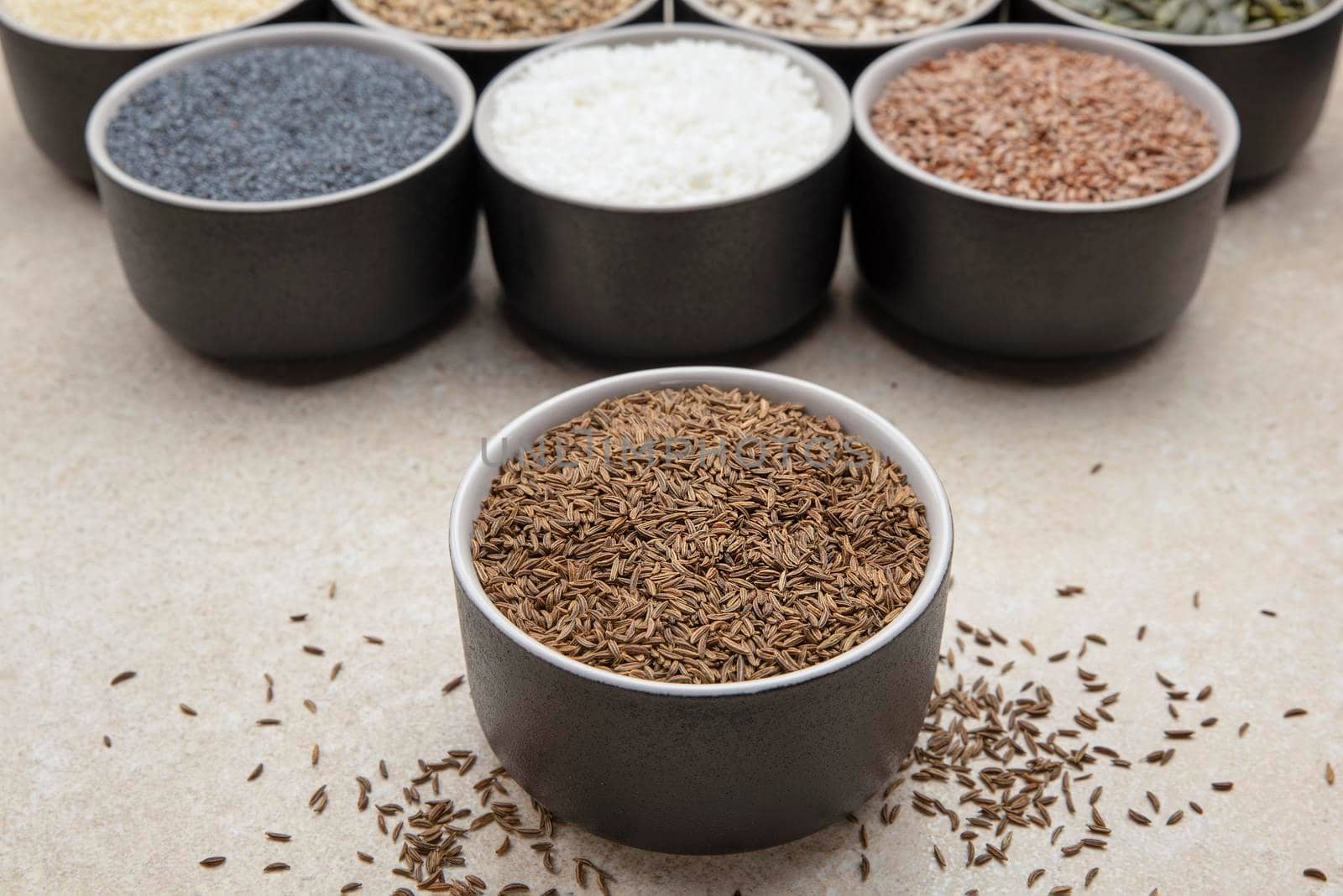 Caraway seeds. Cup with caraway seeds on a background of various seeds in black containers on a marble table top.
