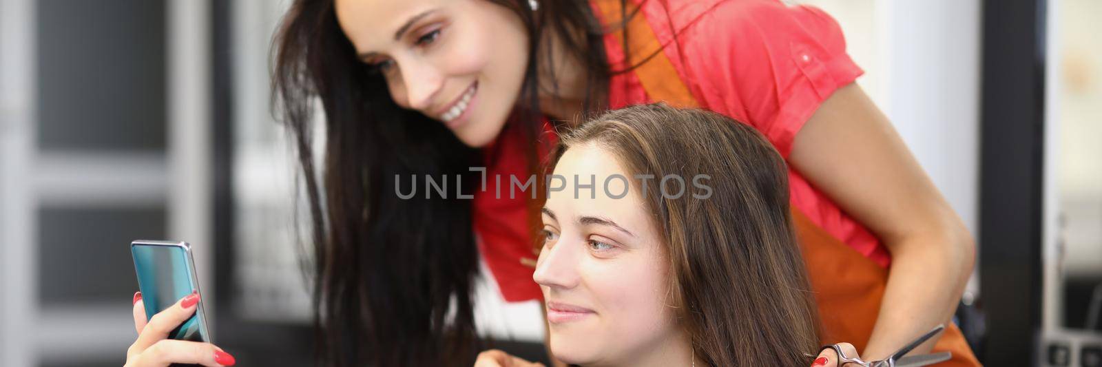 Portrait of smiling client on appointment at hairdresser. Woman show internet content to barber girl on smartphone. Change image, enter new life. Barbershop, style, beauty salon concept