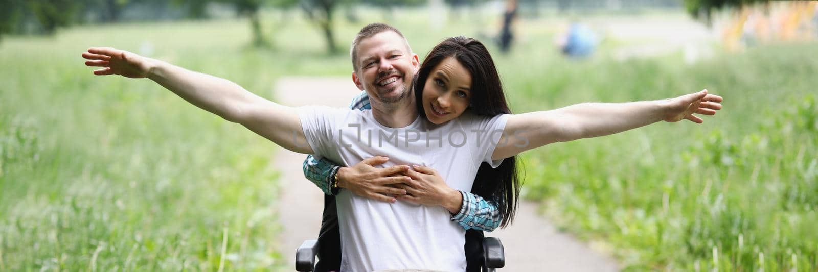Portrait of disabled man sitting in wheelchair, wife hugging from behind. Happy and smiling couple on weekend walk in park. Happiness, appreciate life, accident, love concept