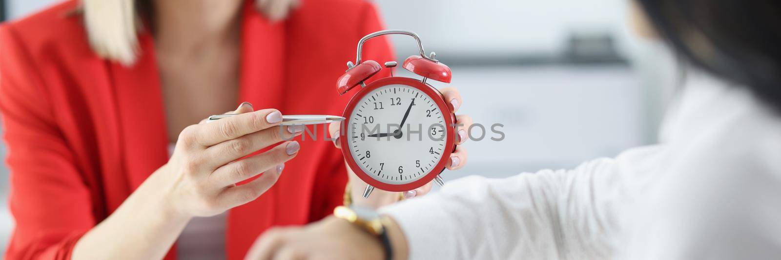 Close-up of boss female berates employee for coming late to work, point with pen at time on red clock. Reprimand from businesswoman to worker, reminder to come on time. Late arrival concept
