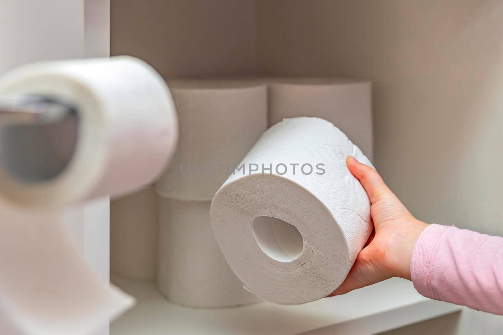 Roll of white toilet paper in hand. The child's hand takes a roll of toilet paper from the cabinet. Lots of toilet paper on background.