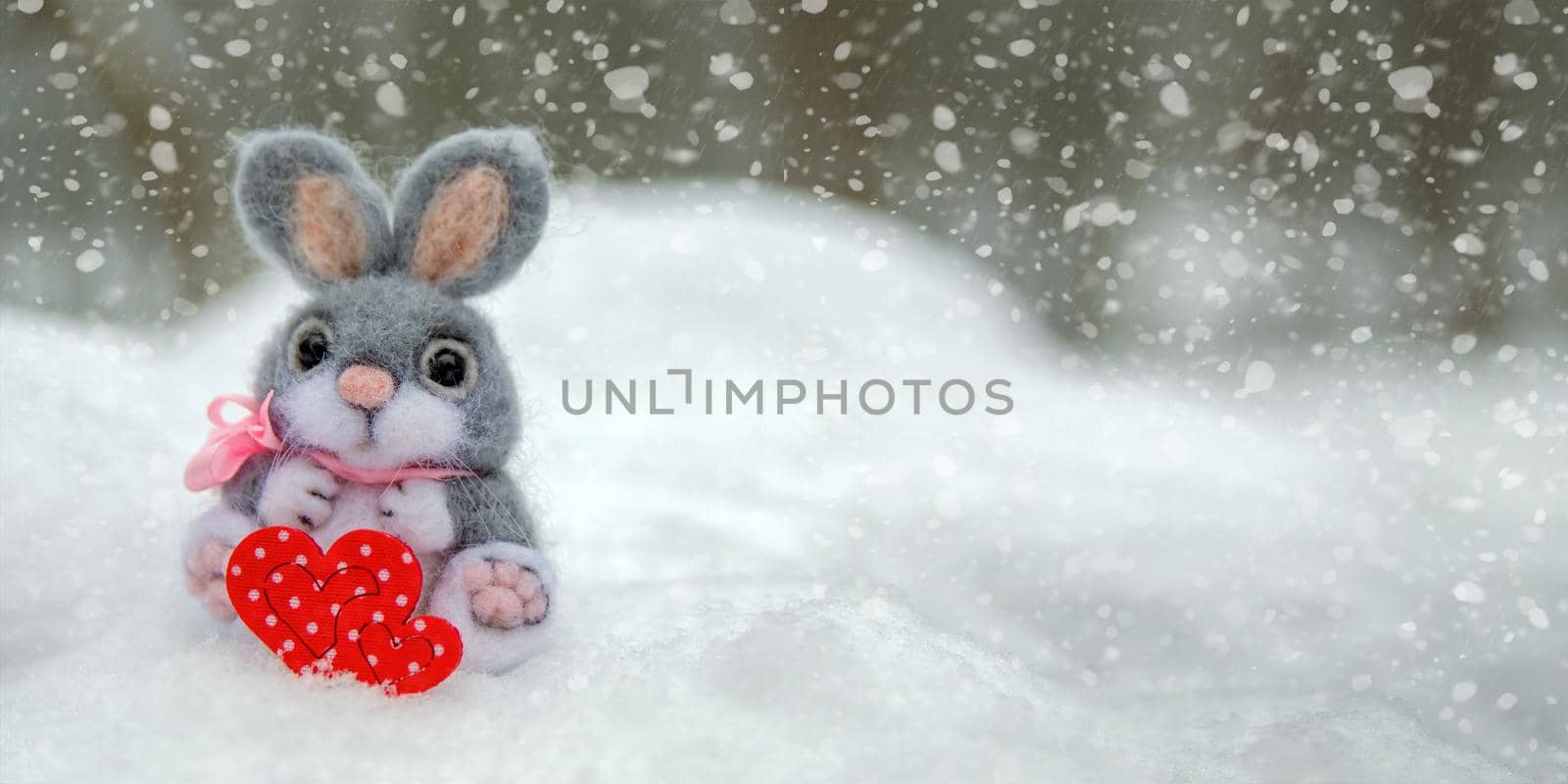 Happy Valentine's Day greeting card or banner. Plush rabbit holding a red heart ka symbol of love. Holidays Happy Valentine's Day. Snowfall on the background of the forest.