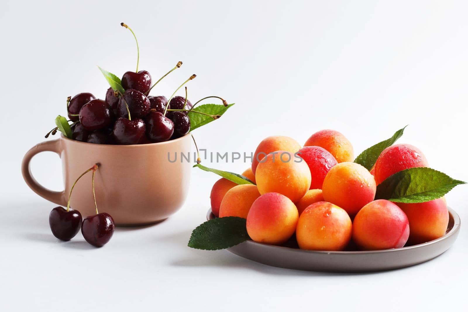 Cherries in a mug and apricots in a plate on a white background. copy space