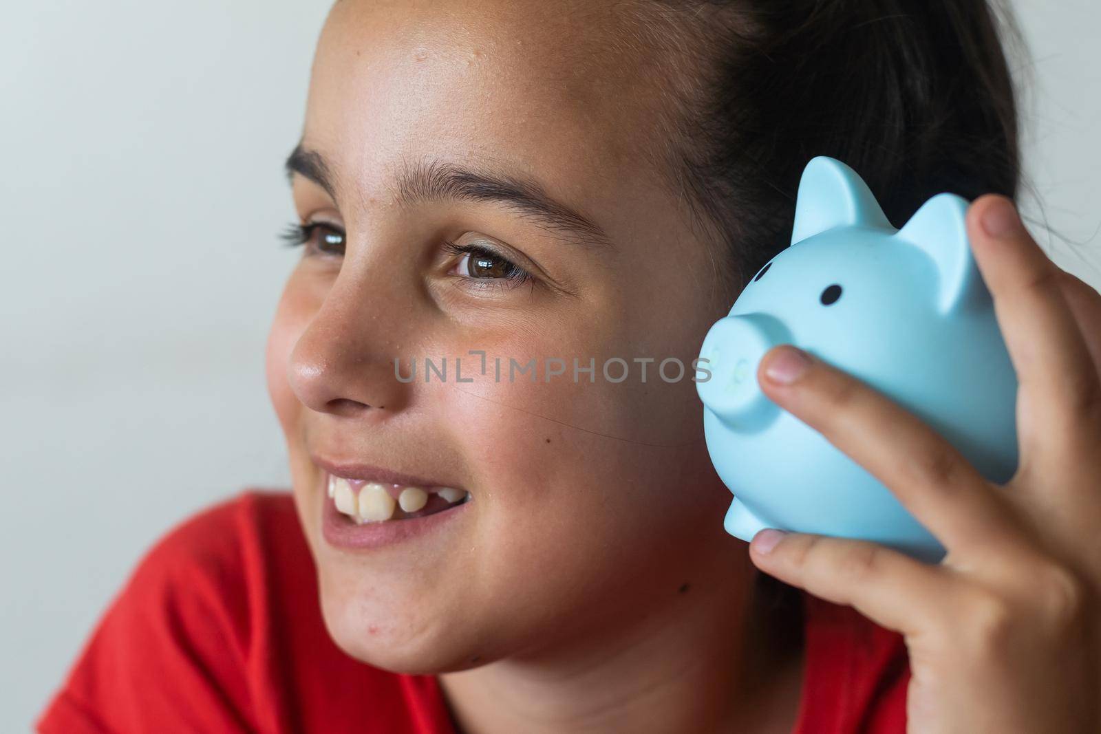 Smiling little girl posing with a piggy bank in her hands, standing against a blue background in a studio with free space. Family savings by Andelov13