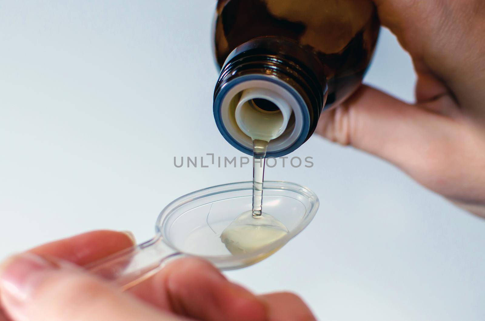Close-up of female hands holding a bottle of expectorant cough syrup and pouring it with a measuring spoon. Health care and treatment concept. Blurred, white background.