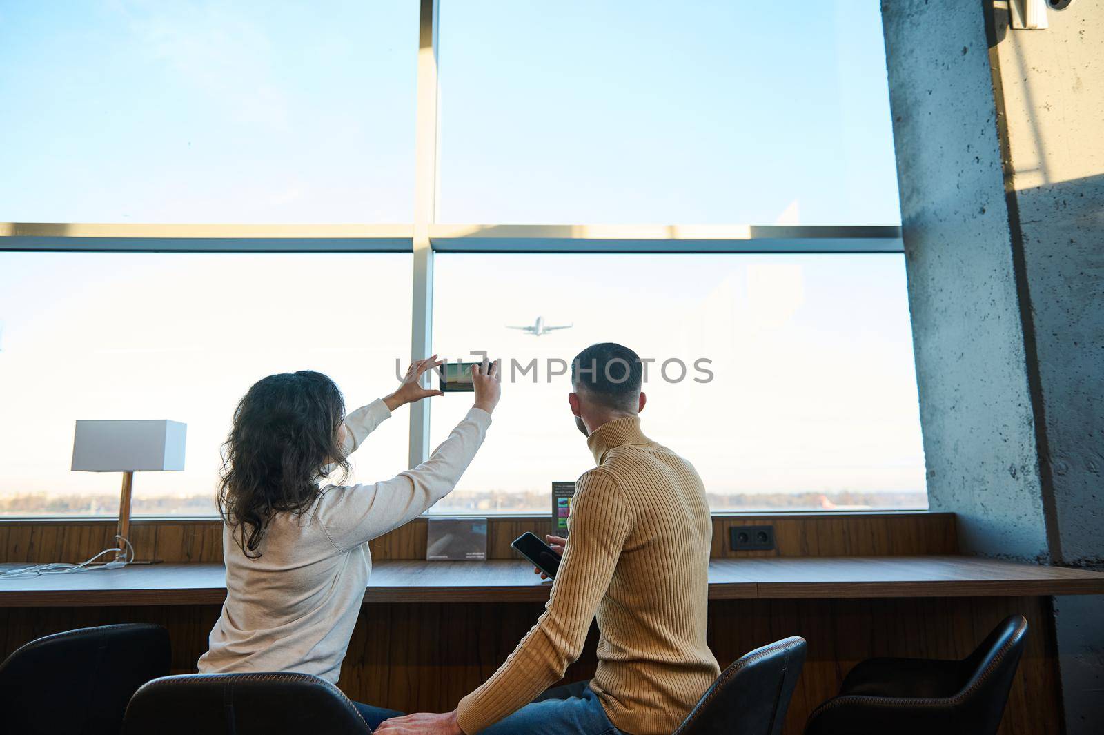 Rear view. Couple of passengers taking pictures of an airplane taking off against beautiful sunset sky, sitting by the panoramic windows in the airport departure terminal, waiting to board the flight