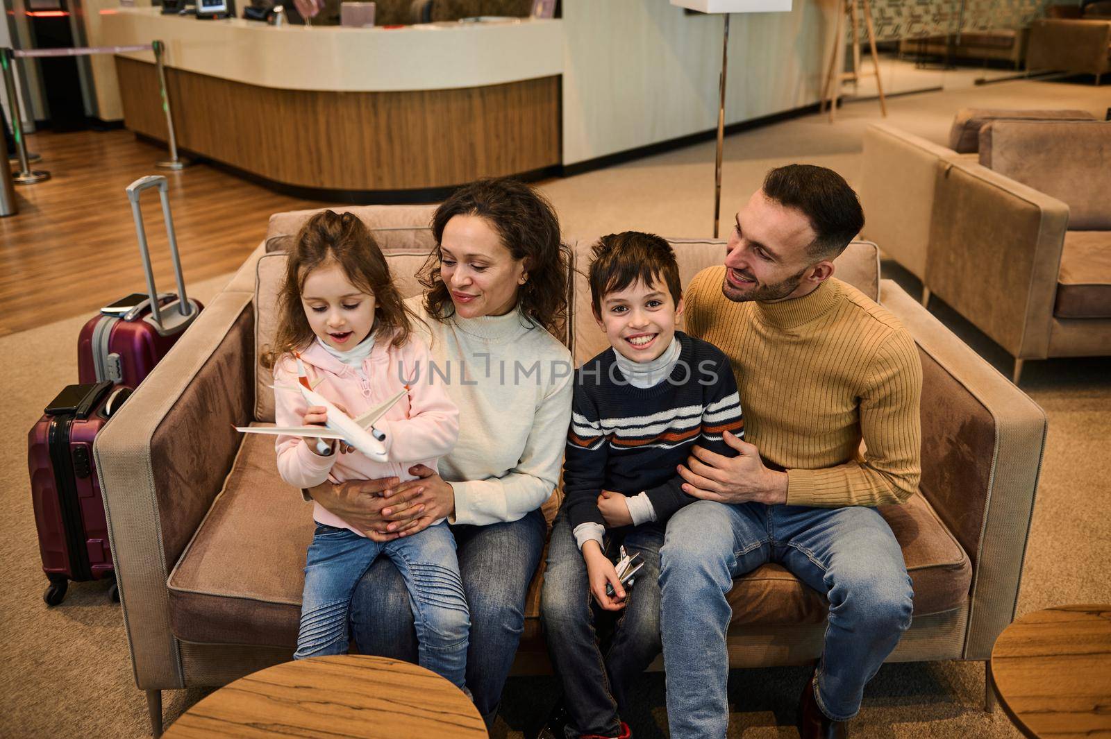 Adorable loving family- mom and dad, gently hugging their kids playing with toy planes while waiting the flight boarding in the VIP lounge area of the international airport departure terminal