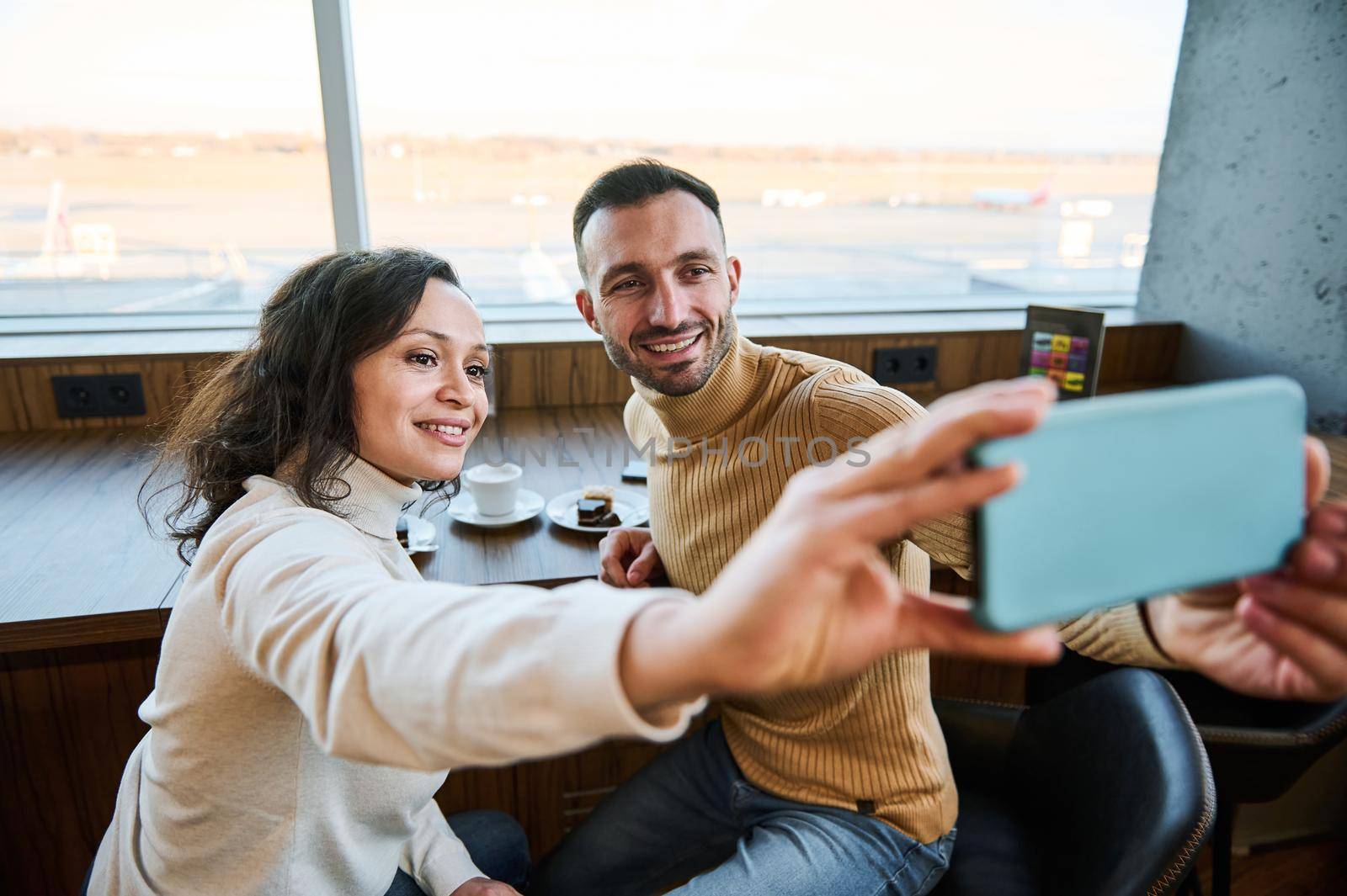 Beautiful multi-ethnic couple in love holding smartphone in outstretched hands and taking self-portrait while enjoying breakfast together in the airport departure terminal before boarding the flight