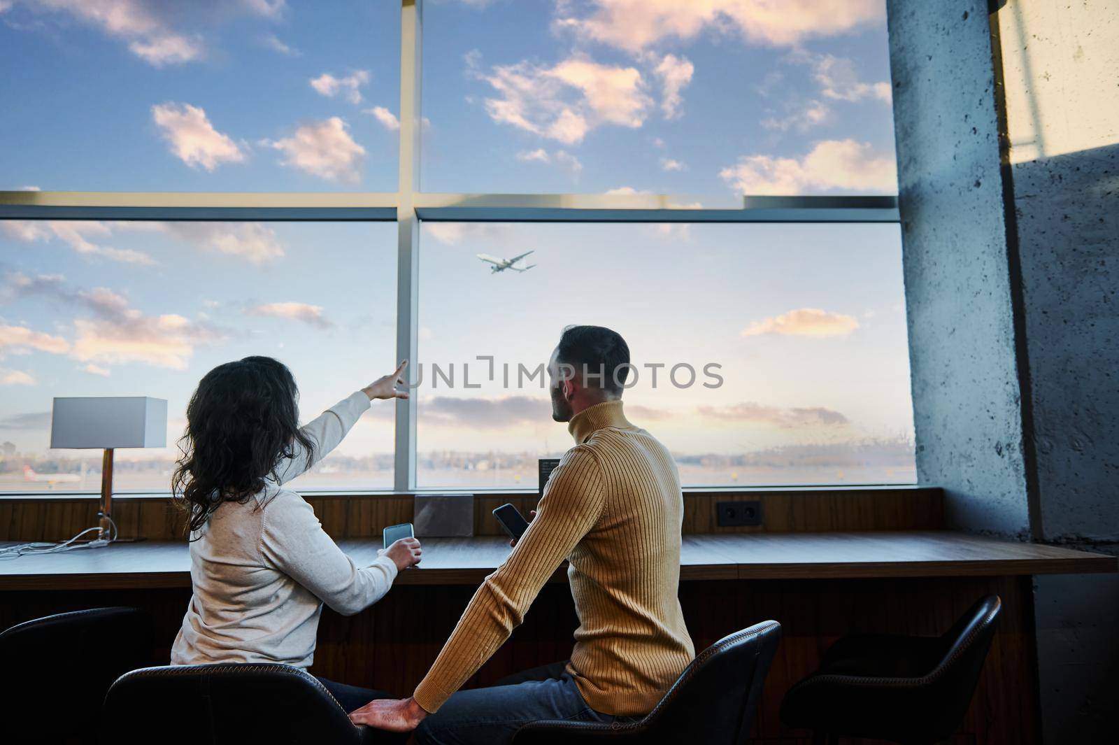 Rear view of a young married couple of passengers admiring a plane taking off, sitting at the panoramic windows in the departure terminal of an international airport, waiting to board the flight