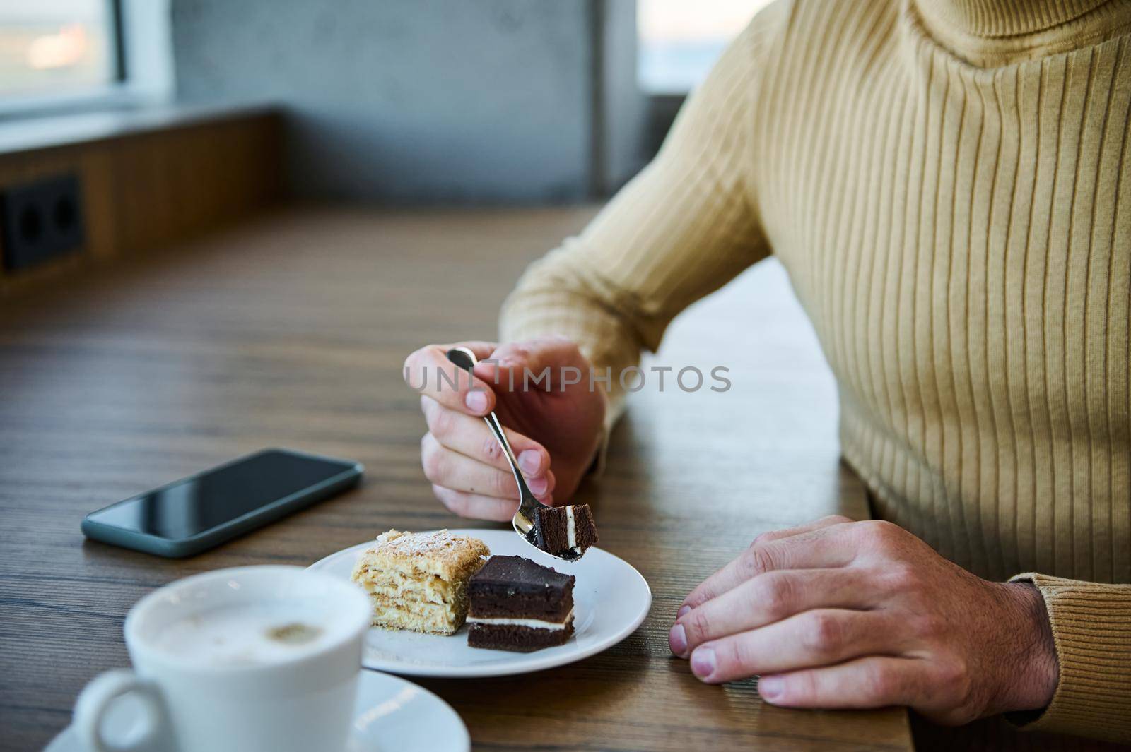 Soft focus on the hands of an unrecognizable man eating chocolate cake and enjoying coffee break at cafeteria. Mobile phone with blank black screen with copy space for advertising text on wooden table