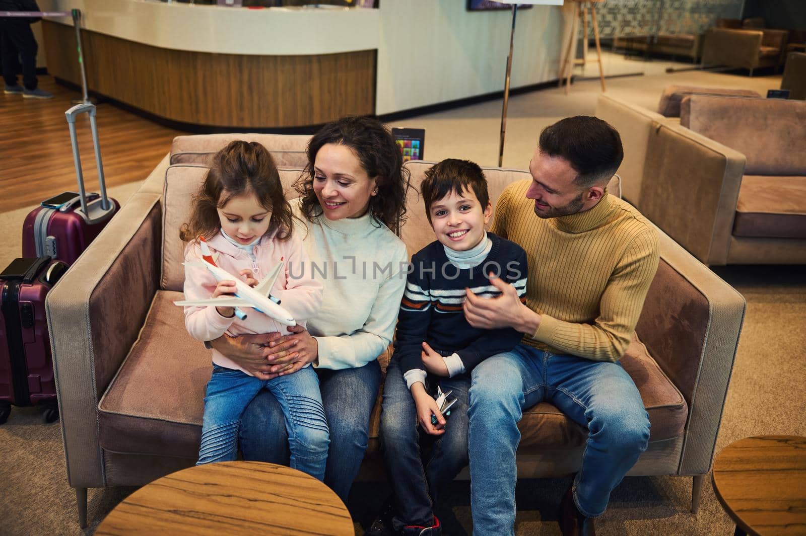 Overhead view of loving mom and dad tenderly hug their son and daughter playing with toy airplanes as they wait to board their flight in the VIP lounge of the international airport departure terminal.