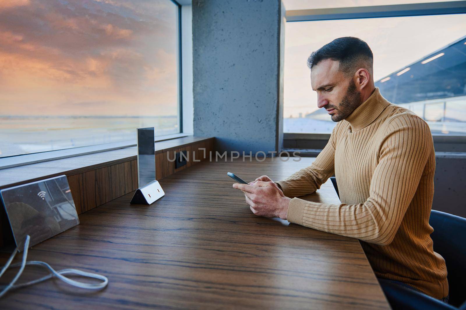 Multitasking man browsing the internet on his smartphone, sitting by windows overlooking runway and beautiful sky at sunset in the VIP lounge of an international airport, waiting to board the flight