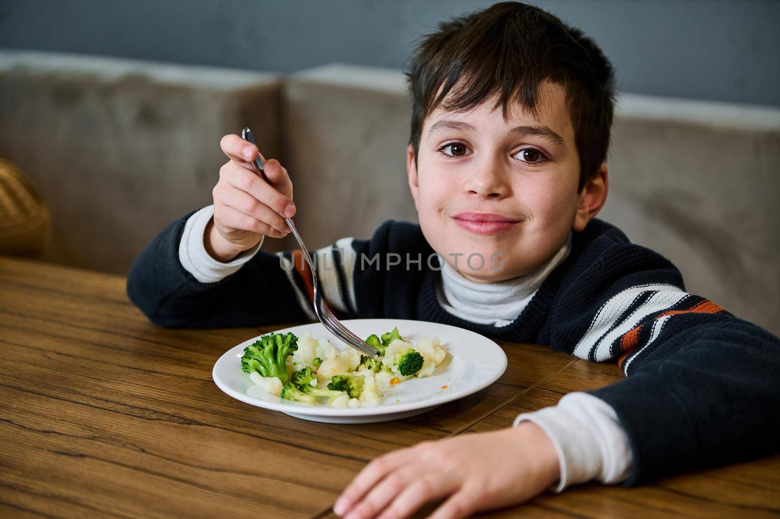 Handsome dark-haired Caucasian school age boy eating healthy vegan meal- steamed vegetables in a canteen and cutely smiling looking at camera