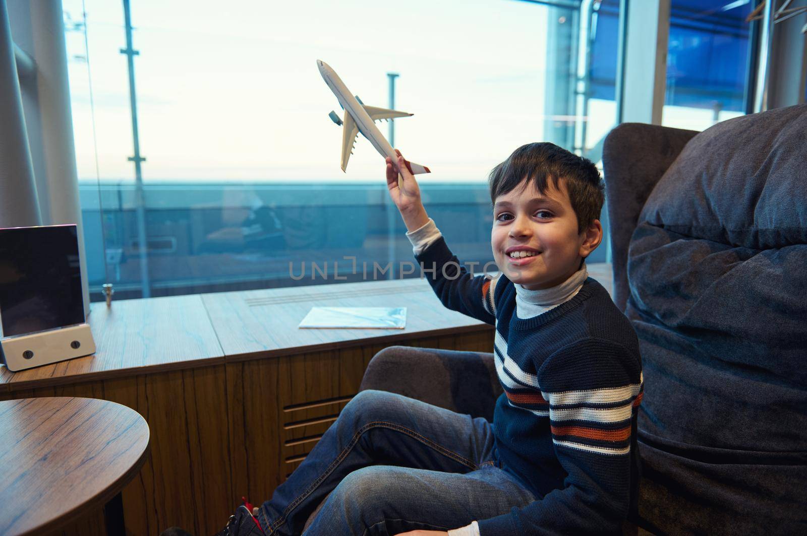 Cute Caucasian boy in warm clothes smiles looking at camera while playing a toy airplane while waiting to board a flight in the VIP lounge of the departure terminal of the international airport