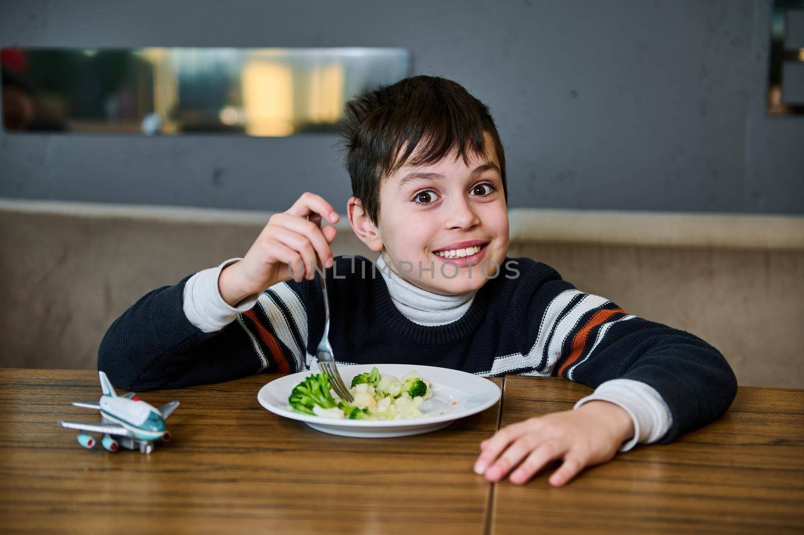 Adorable European boy smiling as he looks at the camera while enjoying a healthy vegan lunch in the canteen of the departure terminal of an international airport. His airplane toy is on a wooden table