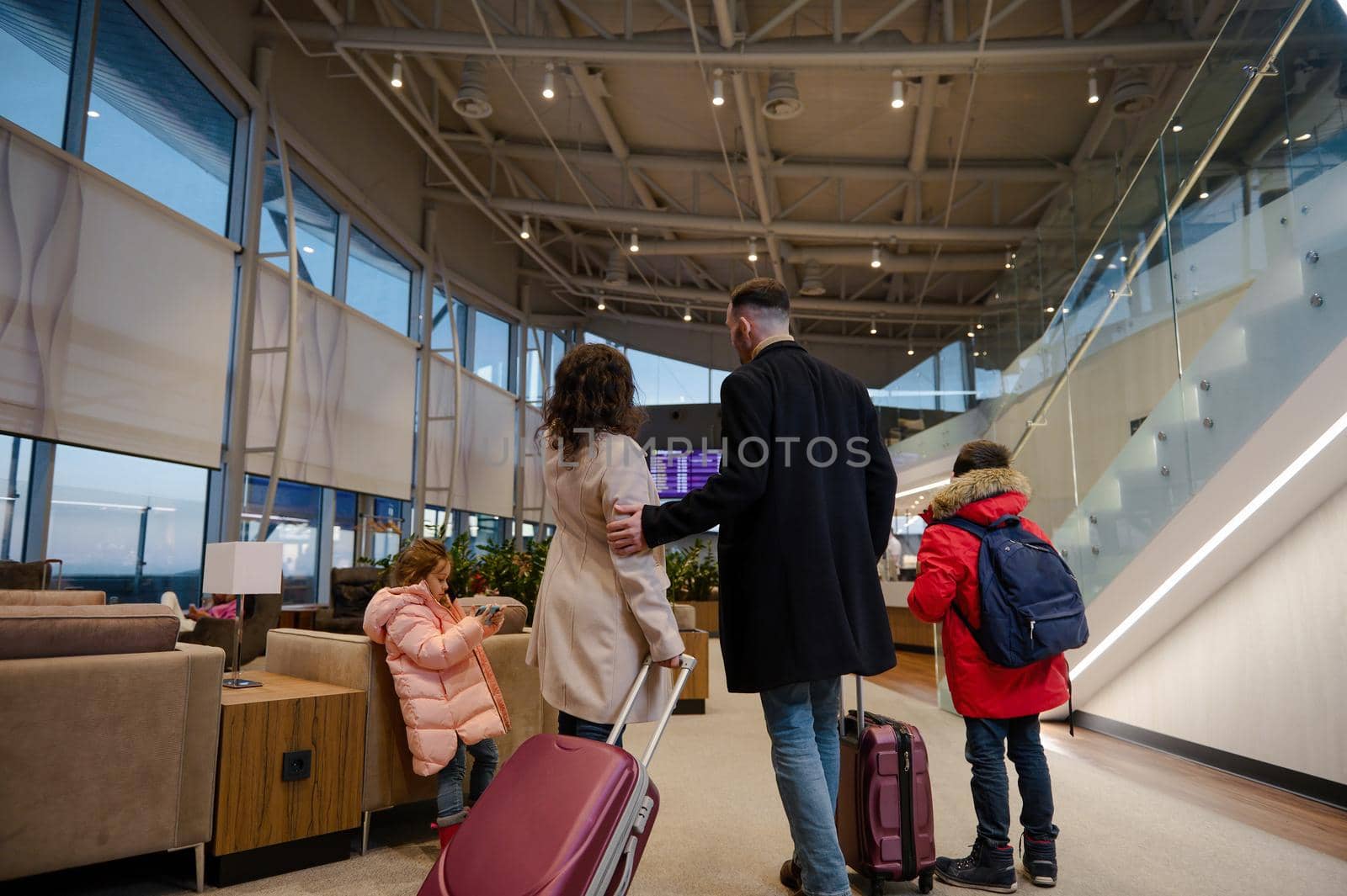 Rear view of traveling family in warm clothes with suitcases checking flight information on board with timetable, standing in the international airport departures terminal, waiting to board flight