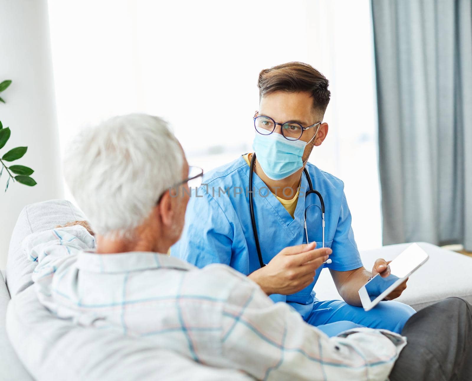 Doctor or nurse caregiver wearing a protective mask helping senior man with a tablet at home or nursing home