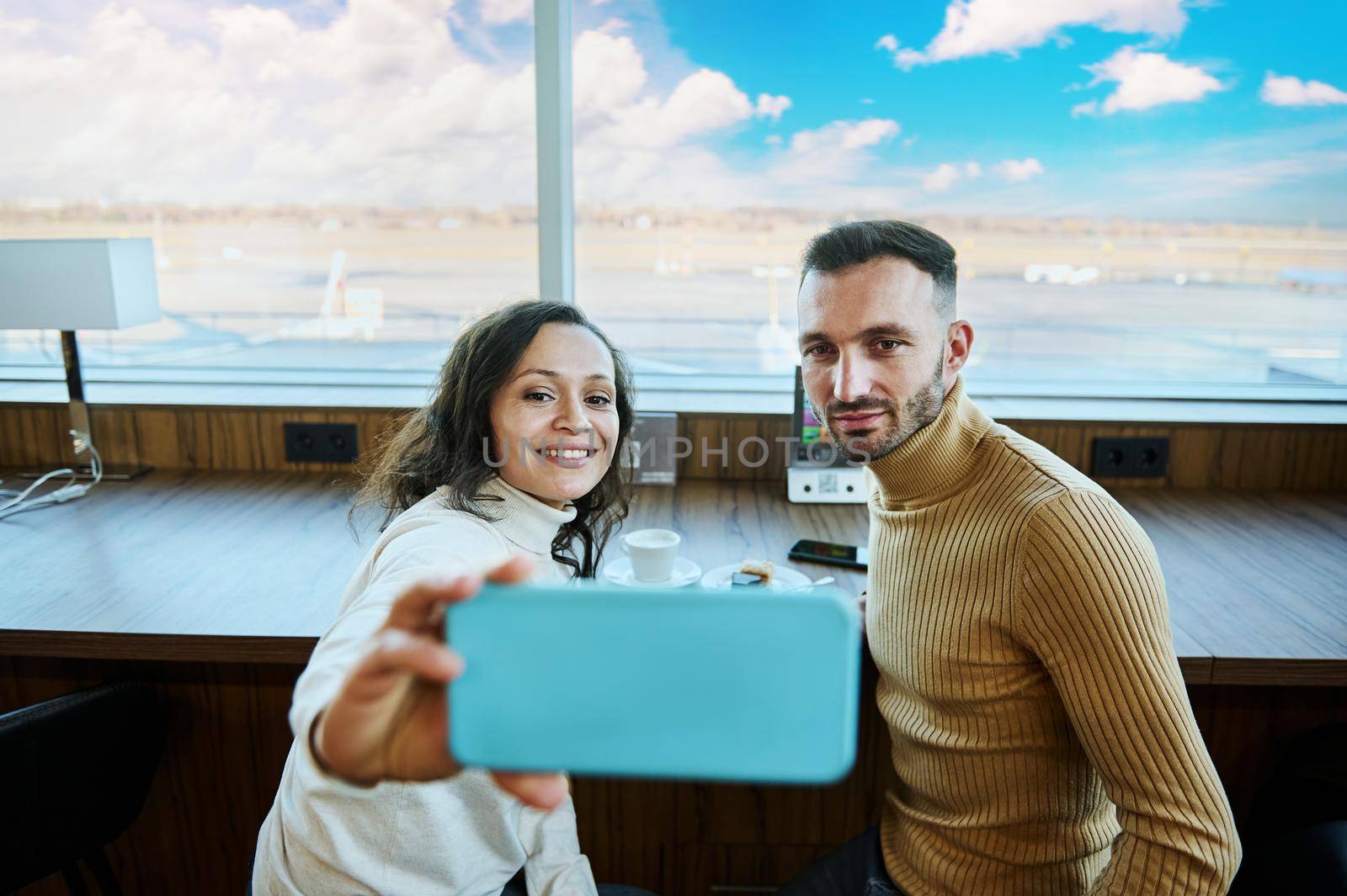 Attractive Middle Eastern young woman sitting next to her husband and taking a selfie against panoramic windows overlooking the runway on a beautiful sunny day while waiting to board a flight