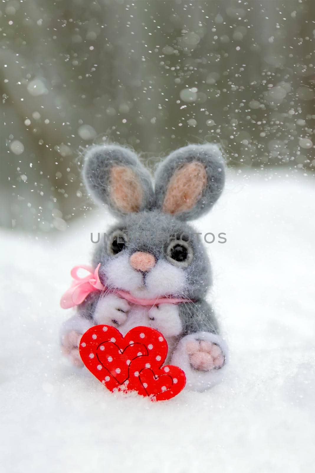 Happy Valentine's Day greeting card or banner. Plush rabbit holding a red heart symbol of love. Holidays Happy Valentine's Day. Snowfall on the background of the forest by SERSOL