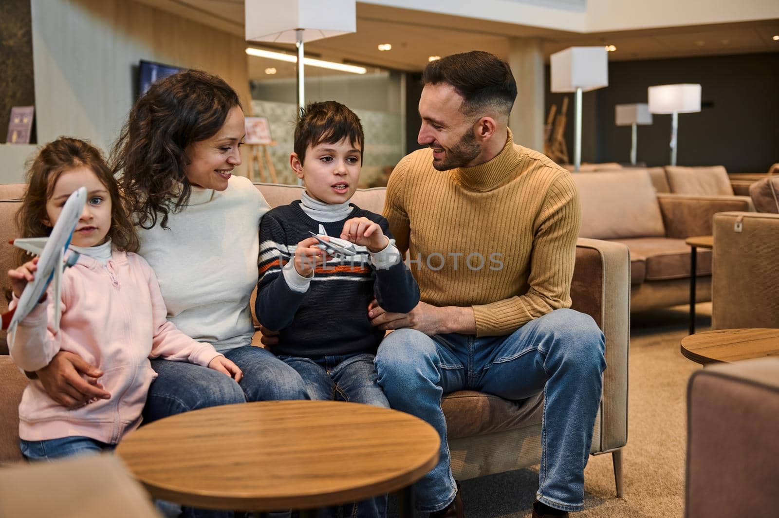 Portrait of happy Caucasian family resting in the VIP lounge of the international airport departure terminal waiting to board the flight. Children - son and daughter playing with toy planes