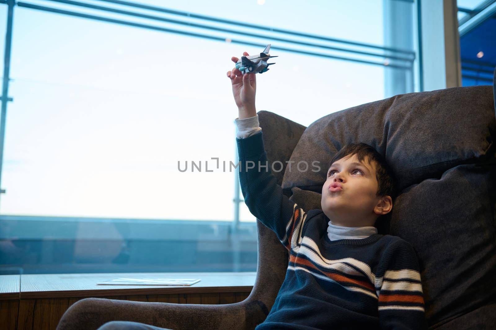 Adorable little boy plays toy plane in VIP lounge of the airport departure terminal sitting on armchair against panoramic windows overlooking runway by artgf