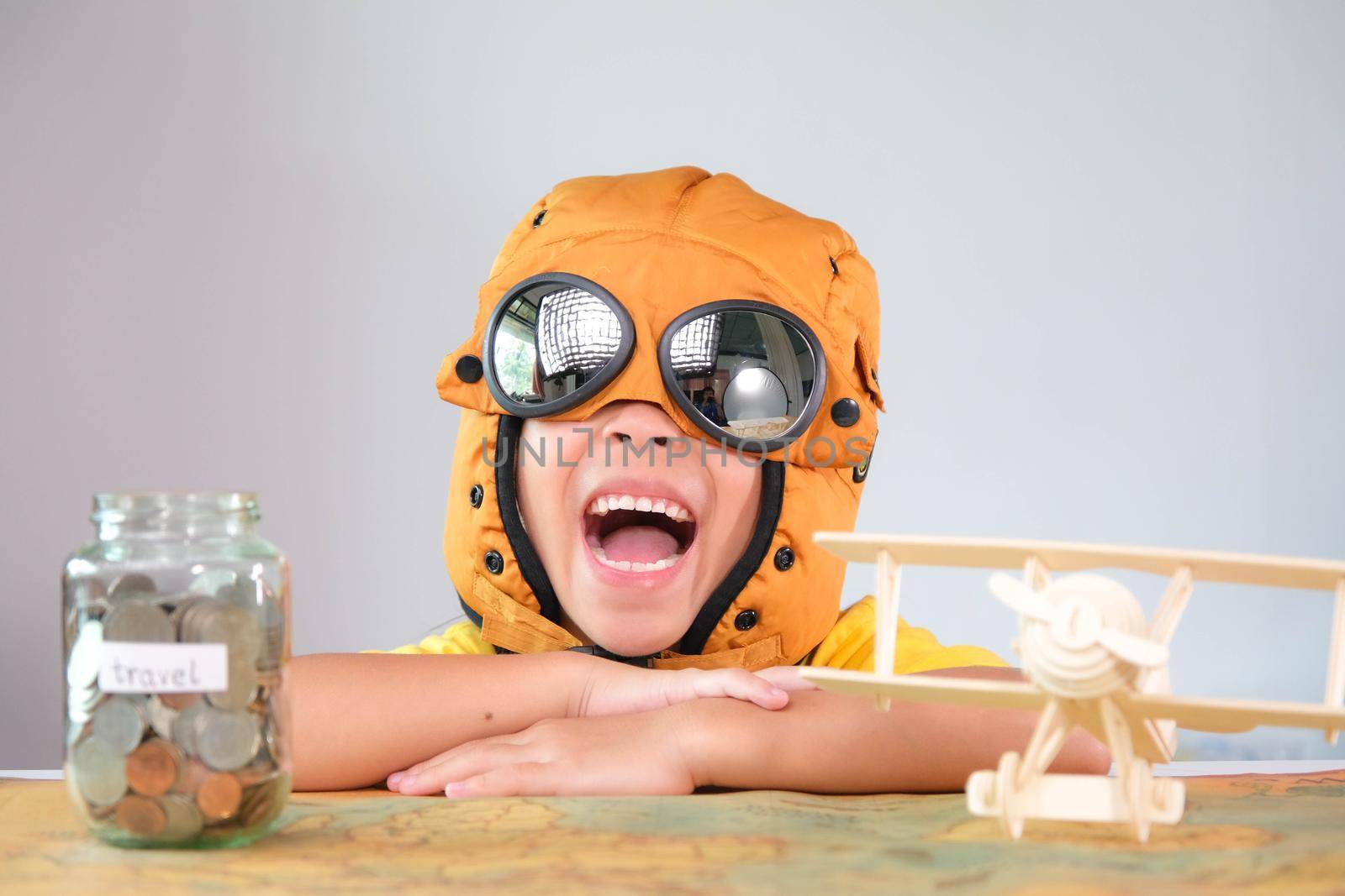Smiling little girl in a pilot hat with coins in a clear jar for savings for travel and a wooden toy plane on the table. Childhood dream imagination and Travel concepts. by TEERASAK