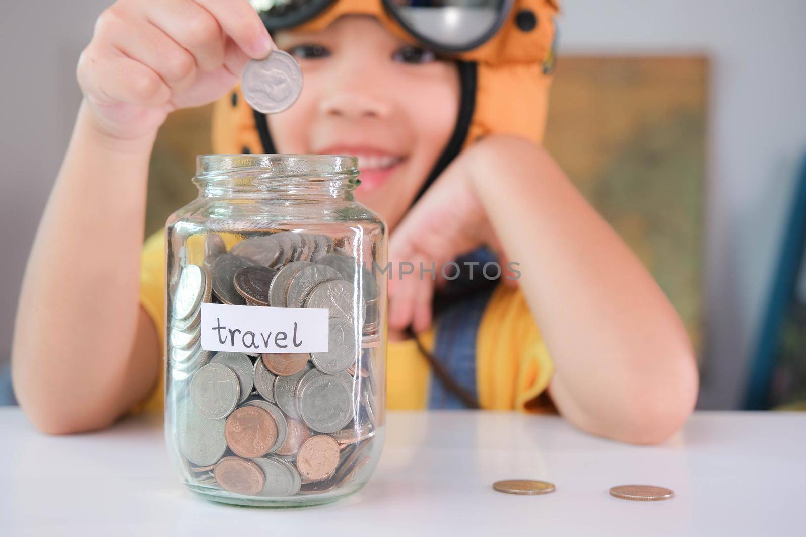 Cute little girl in a pilot hat is putting coins in a clear jar on the table to save for travel. Childhood dream imagination and Travel concepts. by TEERASAK