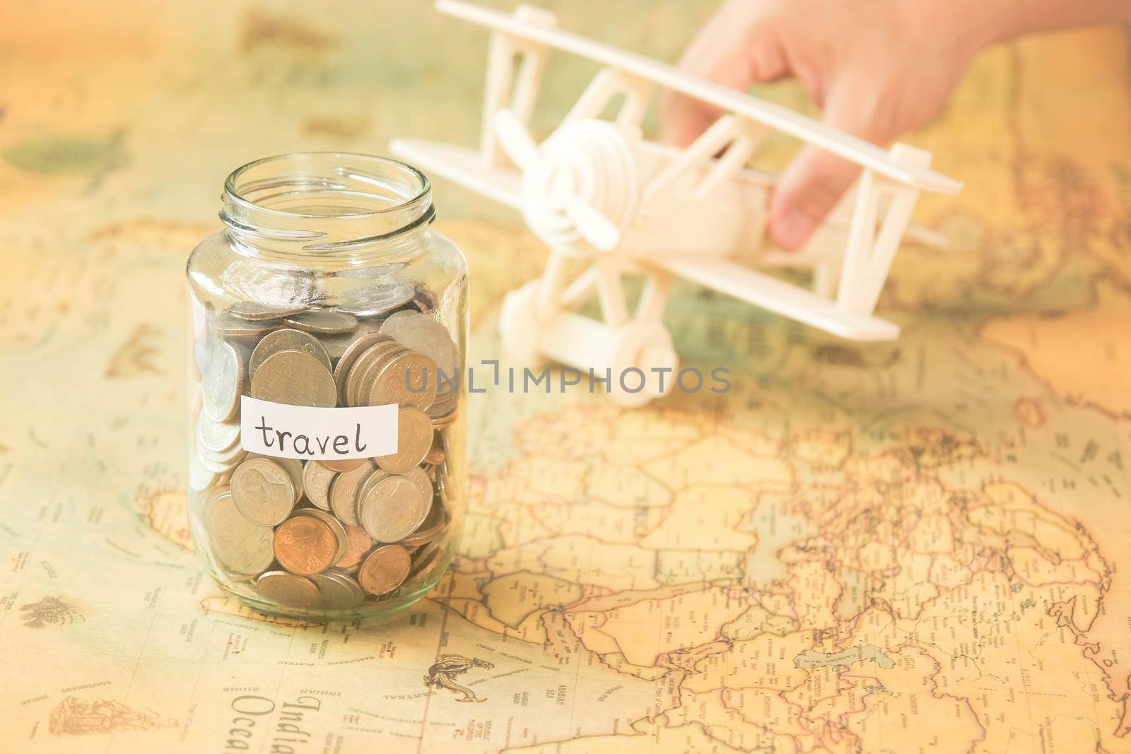 Glass jar with coins and travel inscription on world map with wooden toy plane on table. Travel concept and saving money.