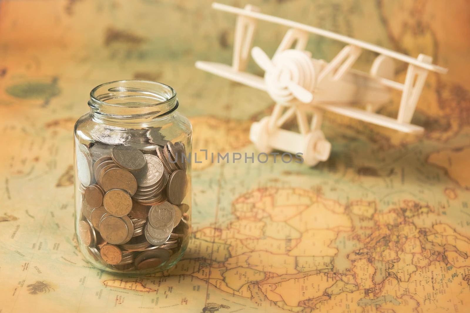 Glass jar with coins on world map with wooden toy plane on table. Travel concept and saving money.