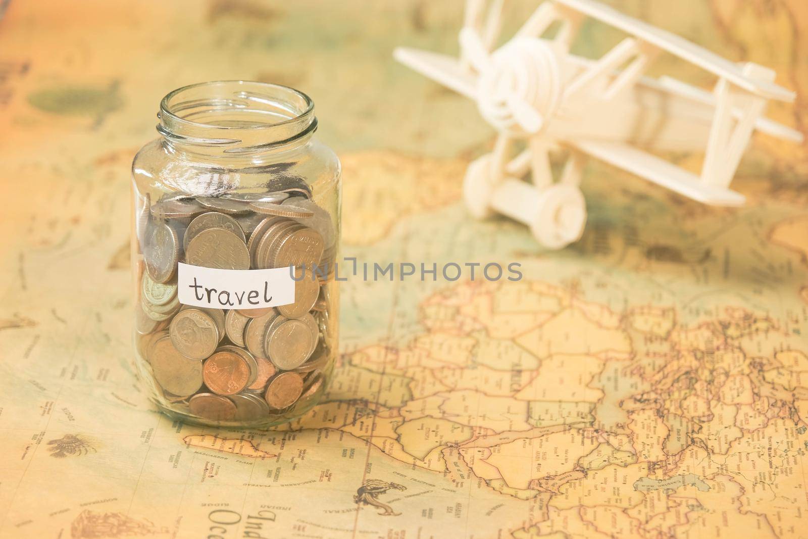 Glass jar with coins and travel inscription on world map with wooden toy plane on table. Travel concept and saving money. by TEERASAK