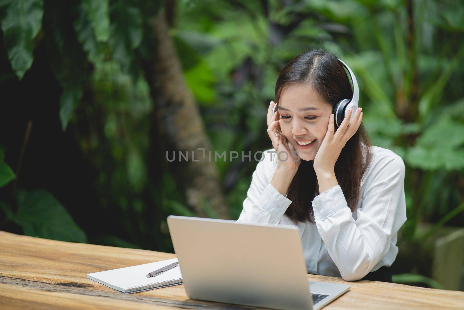 happy young asian woman smile using laptop and wearing white headphone while sitting at cafe. Young asian woman sitting in a coffee shop and video call conference on laptop.