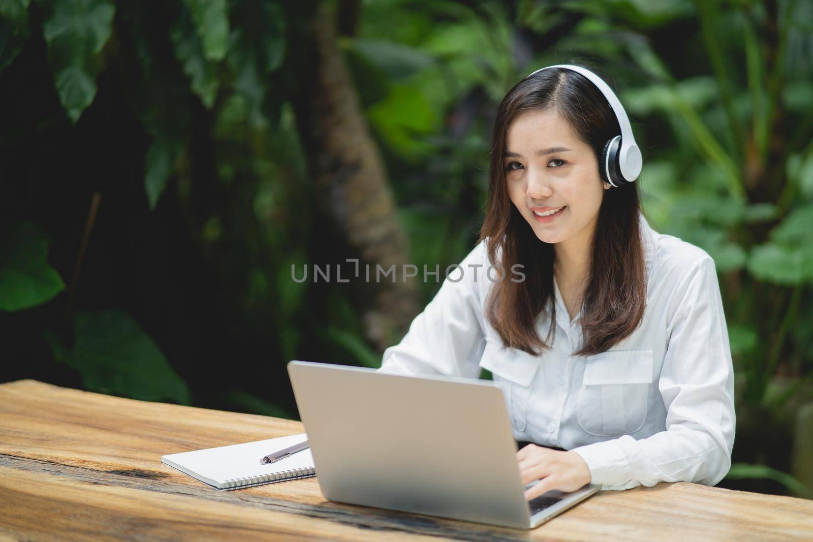 happy young asian woman smile using laptop and wearing white headphone while sitting at cafe. Young asian woman sitting in a coffee shop and video call conference on laptop. by Wmpix