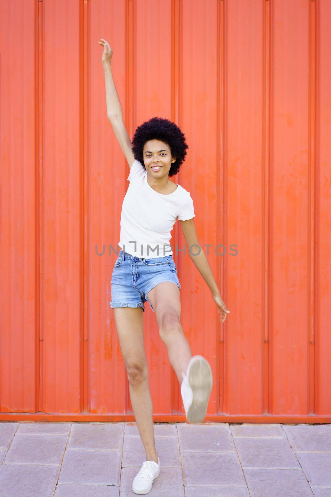 Full body of optimistic African American female in casual clothes looking at camera with raised arm while standing on one leg on red background