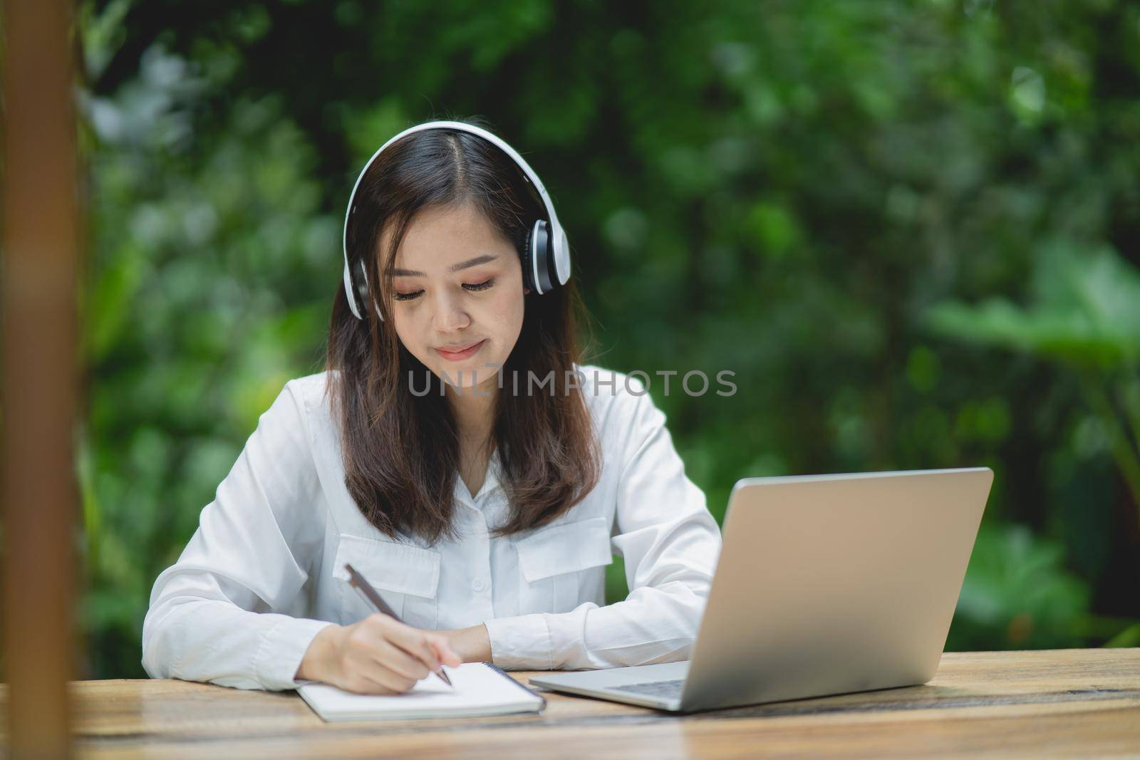 happy young asian woman smile using laptop, wearing white headphone and writing notebook. Young asian woman sitting in a coffee shop and video call conference on laptop. by Wmpix