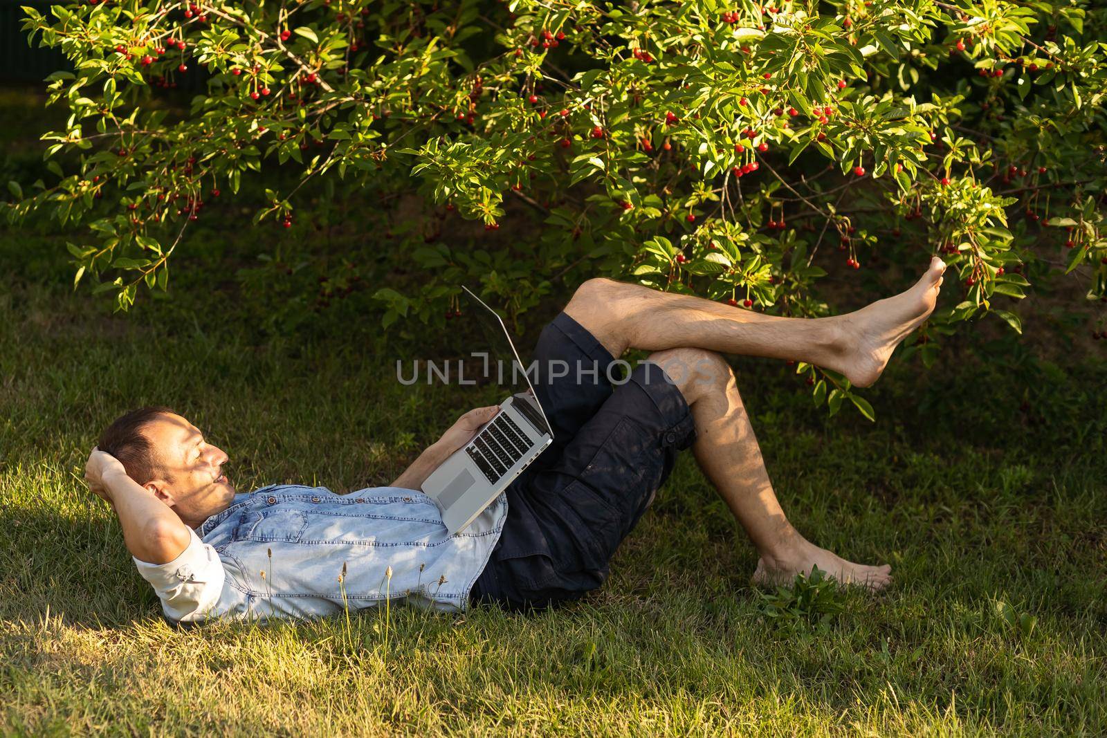 caucasian hispanic man using headphones and sitting on grass working with laptop. Casual man working outdoors by Andelov13