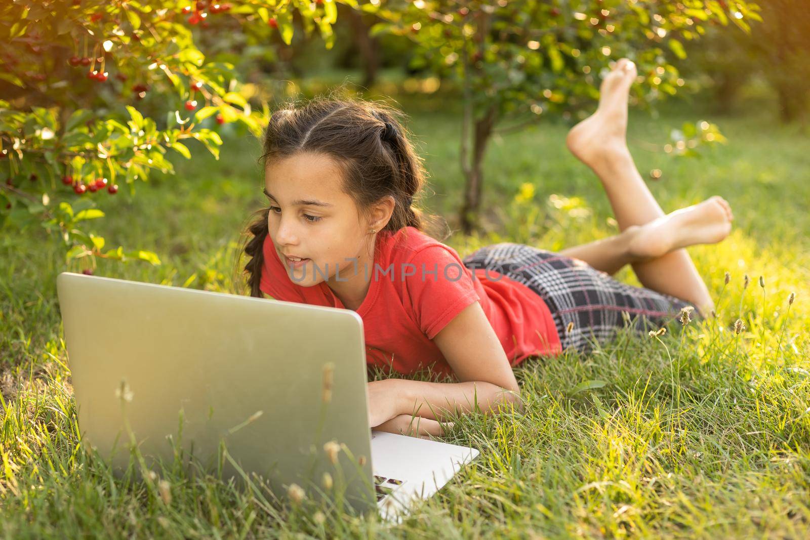 Beautiful young lady is chatting with friends outdoors as it is very useful to be in nature. Laptop is the best friend for a child nowadays. Green and vast meadow.