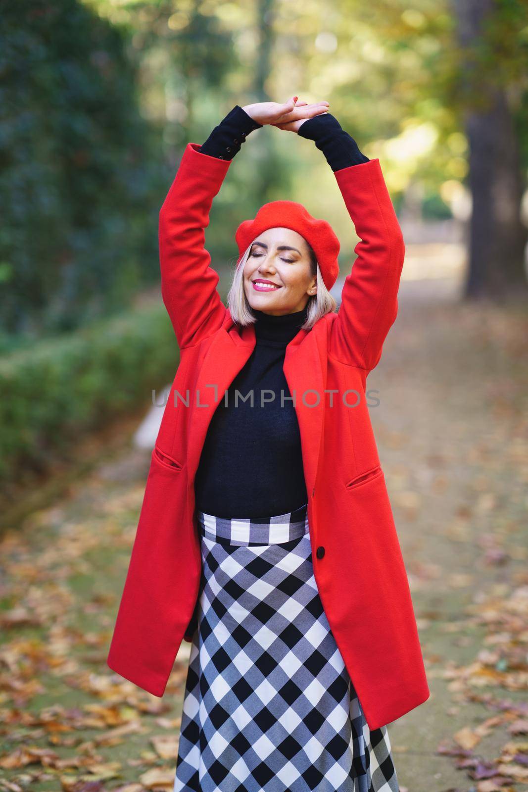 Charming female in stylish red coat and beret standing on path with closed eyes and raised arms in autumn park