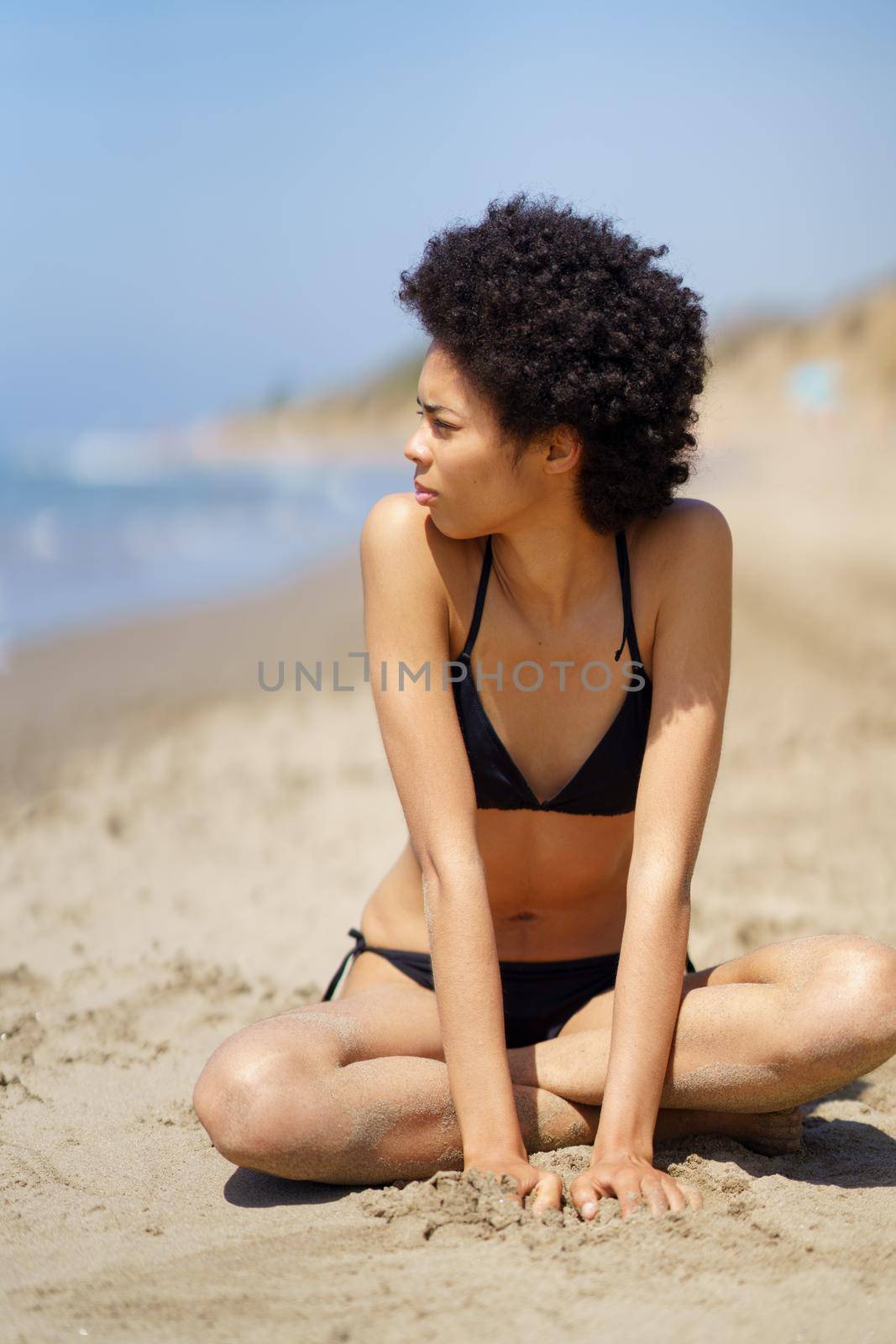 Full body of African American female in swimwear with Afro hair looking into distance while sitting on sandy beach near sea in summer