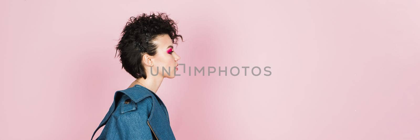 Portrait of a woman with bright flying hair. Beautiful lips and makeup. Hair fluttering in the wind. Web banner. Profile portrait of a young girl in a jeans suit on a pastel pink background.